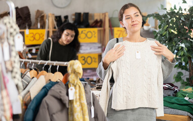 Fototapeta premium European female buyer standing in modern clothing market and carefully examines selection of sweaters on sale of winter clothing collection