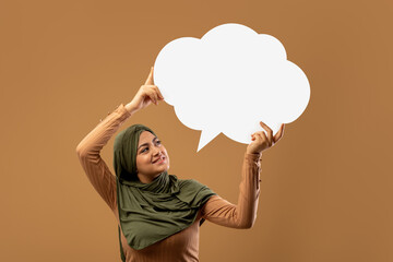 An excited Arab lady stands in front of a brown background, holding a blank speech bubble cloud above her head. She has a joyful expression, indicating an idea or thought.