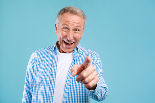 A happy mature man stands in a studio environment with a blue background. He is playfully pointing at the camera, engaging the viewer with a cheerful smile and a friendly demeanor.