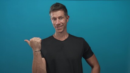 Young man in black t shirt pointing thumb over shoulder showing bare hand gesture in teal studio; confident casual approachable. - Powered by Adobe