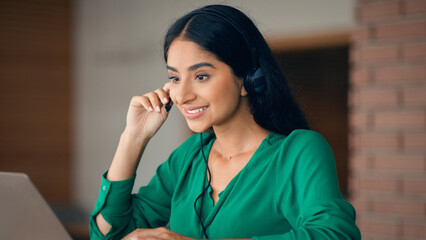 A young woman smiles as she engages in a conversation using a headset while sitting at her laptop in a well-lit office environment. She appears focused and attentive during her work.