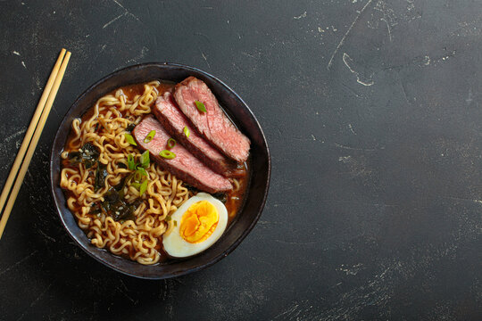 Bowl of ramen noodle soup with sliced beef, a boiled egg, seaweed, and green onions, served with chopsticks on a dark background, asian style soup top view, copy space