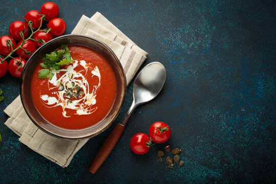 Fresh tomato soup is served in a bowl with cream, seeds and herbs, accompanied by cherry tomatoes, garlic and parsley on a blue background with a spoon and napkin, copy space - Powered by Adobe