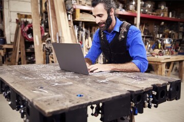 Craftsman in the Workshop: A skilled craftsman, engrossed in his work, meticulously operates a laptop at his workbench, surrounded by the tools of his trade.