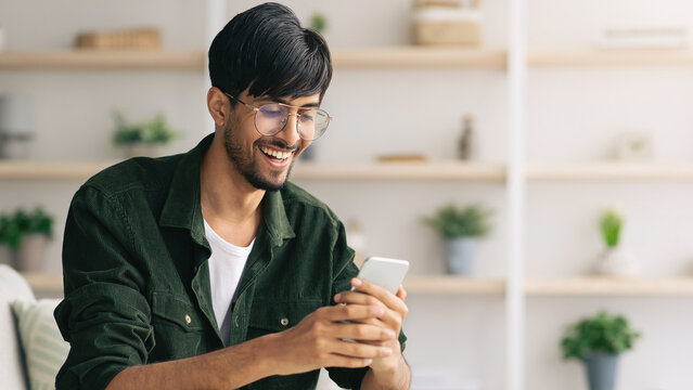 A young man laughs while focused on his smartphone in a bright, modern living space. Plants and decor enhance the warm atmosphere of the room. - Powered by Adobe