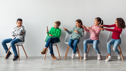 Fototapeta premium Preteen kids are laughing at an African American boy sitting alone in a classroom. The boy is focused on his smartphone while others point and giggle, highlighting bullying and social issues.