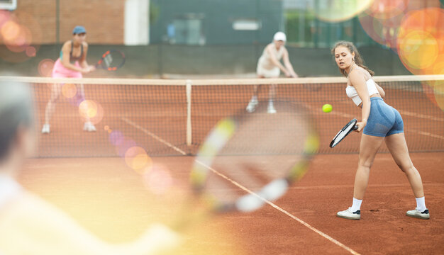 Emotional fit young girl, seen from behind, enthusiastically playing tennis outdoors, performing left-handed forehand to return ball to opponent court during doubles match with female partner..