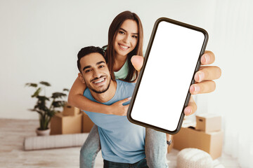 A young couple shares a joyful moment indoors with the man giving the woman a piggyback ride. They smile at the camera while the man holds up a smartphone, ready to capture fun memories.