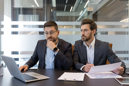Two professional businessmen are analyzing data on a laptop and reviewing documents, collaborating on a project during a corporate discussion in a modern office meeting room