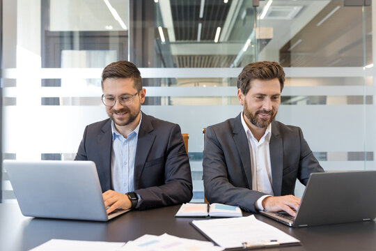 Two smiling bearded businessmen in suits working on individual laptops at a bright modern office desk, focused on planning, collaboration and productive teamwork tasks - Powered by Adobe