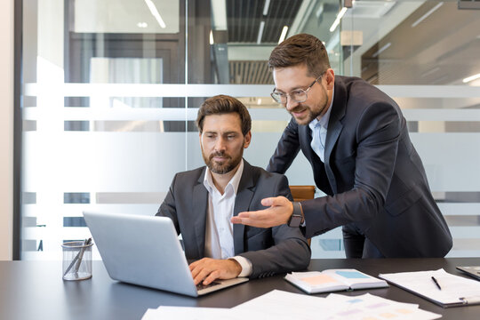 Two male businessmen in suits discussing business strategy and reviewing data on a laptop, working together on a project in a modern office environment