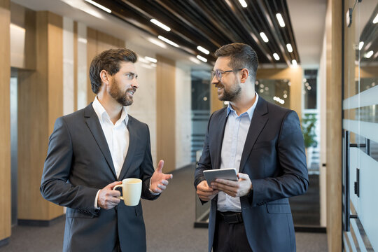 Two professional businessmen discussing a new project, standing in a contemporary office corridor, one holding a coffee cup and the other a digital tablet while exchanging ideas - Powered by Adobe
