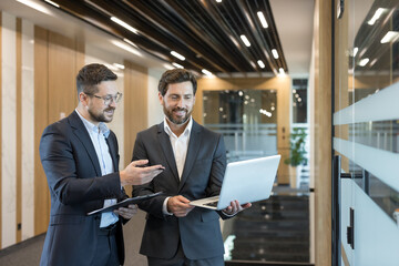 Two professional businessmen collaborating and discussing a corporate project, one holding a laptop while the other points to a clipboard, standing in a modern office hallway