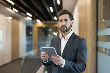 Fototapeta premium Confident bearded businessman standing in a contemporary office hallway, holding a digital tablet, embodying professionalism, technology, and success in a corporate setting