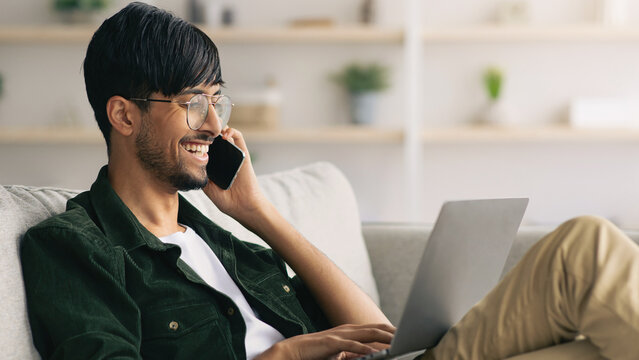 A young man sits on a comfortable couch in a bright living room, smiling while talking on his phone. He types on a laptop resting on his lap, enjoying a productive moment at home.