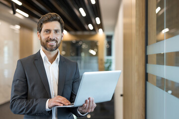 Confident businessman standing and smiling. Working on a laptop while dressed in a stylish suit in a contemporary office hallway. Representing professional success. Technology. And business management