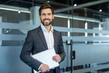 Confident businessman smiling at camera, standing in a contemporary office environment, holding a...