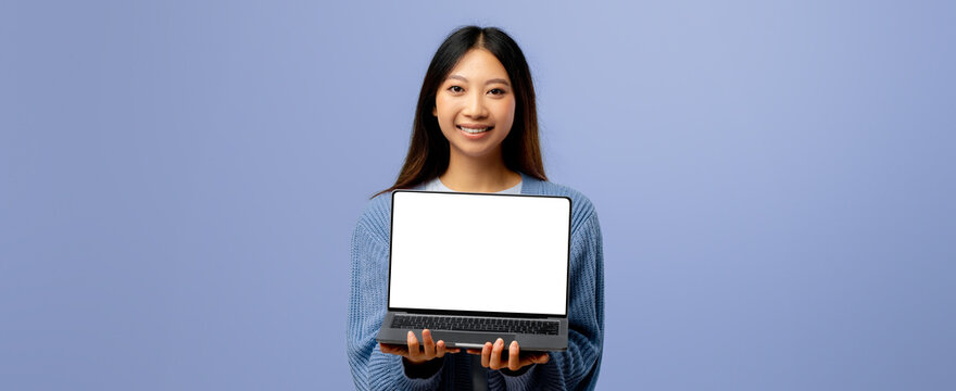 A cheerful young woman stands proudly with a laptop in her hands. She is dressed in a cozy sweater and poses in front of a light blue background.