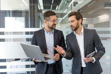 Two male business colleagues discussing project strategy and innovative ideas while looking at a laptop computer, standing together in a modern corporate office environment