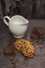 Milk jug and oatmeal cookies on the table. Pieces of broken dark chocolate. Spilled coffee beans. Dark background. Coffee service. Still life