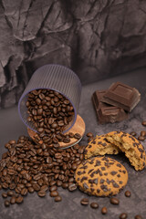 Coffee beans spill out of a transparent glass, oatmeal cookies on the table. Pieces of broken dark chocolate. Dark background. Still life