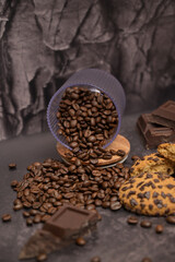 Close up. Coffee beans spill out of a transparent glass, oatmeal cookies on the table. Pieces of broken dark chocolate. Dark background. Still life