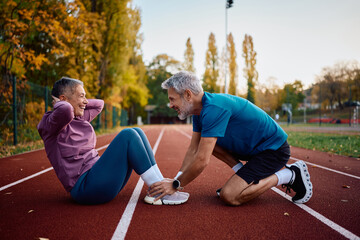 Fototapeta premium Happy mature athlete assisting his wife in doing sit-ups during outdoor workout in autumn.