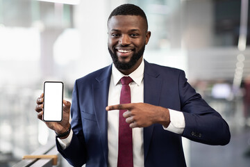 Positive young black businessman pointing at brand new smartphone with white empty screen, recommending newest mobile application for business online, office interior, mockup, copy space