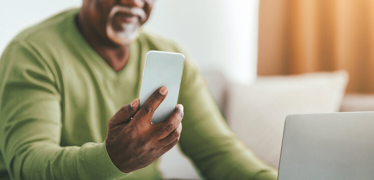 A man in a green sweater sits comfortably at home, engaging with his smartphone while his laptop is open next to him. Sunlight filters softly through a nearby window.