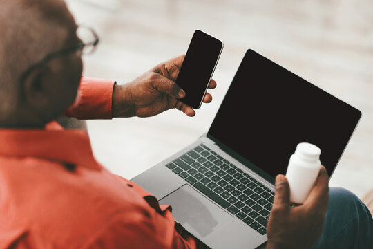 An elderly man wearing glasses sits comfortably, holding a smartphone in one hand and a medication bottle in the other, with a laptop open in front of him.
