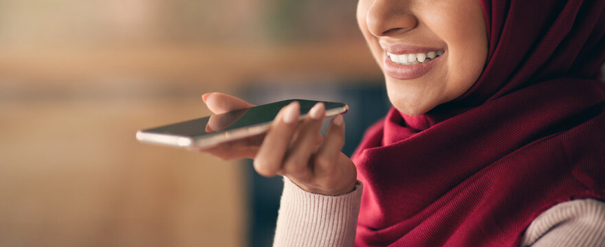 A young woman with a red hijab smiles while holding her smartphone. She appears engaged in a conversation, enjoying the ease of using voice commands in a warm, inviting environment.