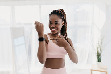 A young black woman enjoys a break in her home workout. She smiles while pointing at her fitness tracker. Natural light enhances the positive atmosphere in the living room.