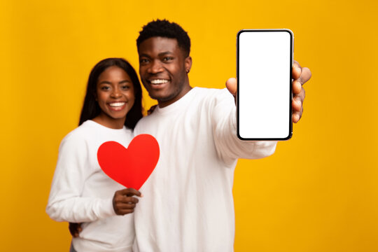 A happy couple stands together, smiling and holding a red heart and a blank phone. They wear matching white shirts and pose in front of a vibrant yellow background, creating a cheerful atmosphere.