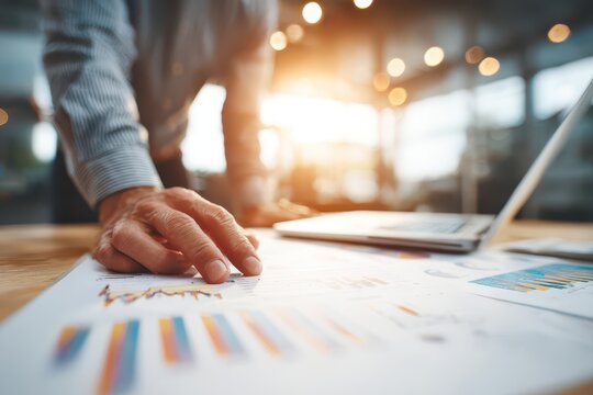Close-up of a businessman's hand analyzing financial data charts on a desk with a laptop and bright light. - Powered by Adobe