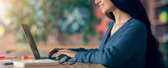 A woman sits at a wooden table in a well-lit cafe, focused on her laptop. She smiles while typing,...