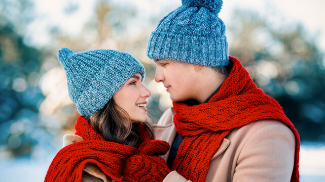 A young couple stands close, smiling at each other in a snowy landscape. They are dressed warmly in knit hats and red scarves, enjoying a cozy moment in the winter sun.