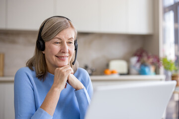 Happy senior woman with gray hair wearing a headset, smiling and working on a laptop in her bright home kitchen during a video call or remote work session