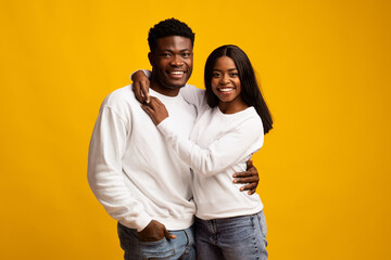 A happy couple stands close together, embracing each other with smiles. They wear matching casual outfits, creating a cheerful and loving atmosphere against a bright yellow backdrop.