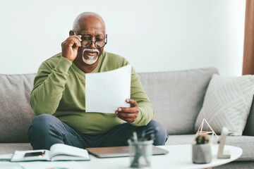 A joyful senior man sits comfortably on a couch at home, examining papers with a smile. He wears...