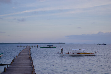 Fototapeta premium A serene wooden pier stretches over a calm ocean, with traditional Philippines boats floating nearby, embraced by a tranquil setting under a gentle sunset sky.