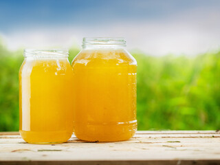 Two glass jars of golden bee honey are on a wet table, green grass field and sky in the background. Fine organic product concept. Agriculture and food industry.