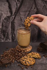 Woman's hand dips a piece of oatmeal cookie into coffee cappuccino latte in a transparent glass on the table. Pieces of broken dark chocolate. Spilled coffee beans. Dark background. Coffee service