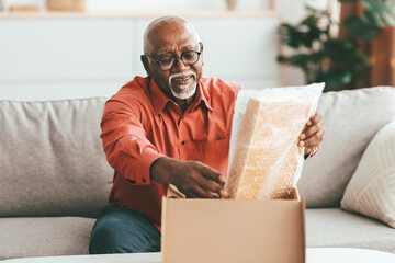 An elderly man sits on a sofa in his living room, excitedly unboxing a package filled with bubble wrap. He shows joy as he discovers the contents of the box in the afternoon light.