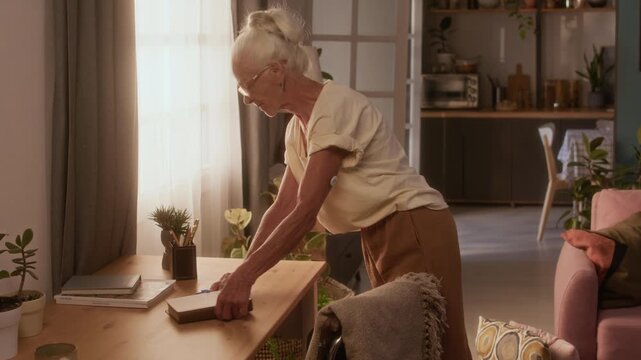 Medium shot of elderly Caucasian lady, wearing short sleeve blouse and adhesive diabetes patch on upper arm cleaning up dust on table with cloth, while doing household chores at daytime