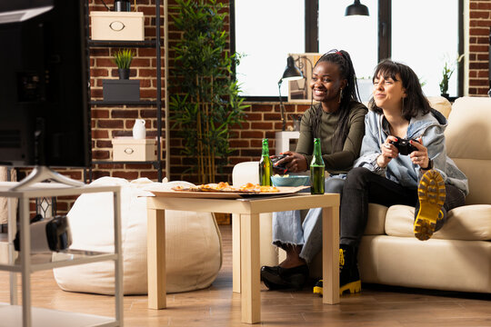 Diverse female friends using wireless controllers, sitting on couch with excitement while playing video games. Happy black and white women enjoying online gaming during fun weekend hangout.