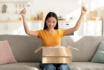 An excited Asian woman sits on a couch in her bright living room, joyfully unpacking a delivery box. She clutches her fists in happiness, revealing her delight about the surprise package.