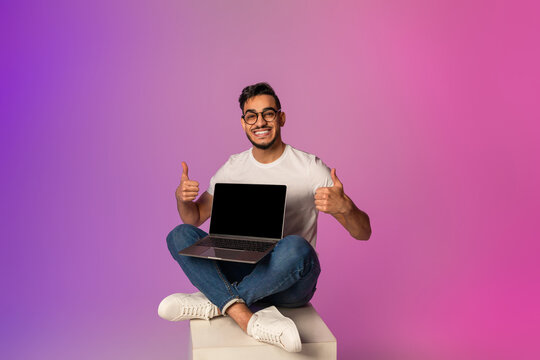 Cheerful young Arab man sits confidently with a laptop featuring an empty screen. He gestures thumbs up, enthusiastically recommending a new website against a vibrant neon backdrop. - Powered by Adobe
