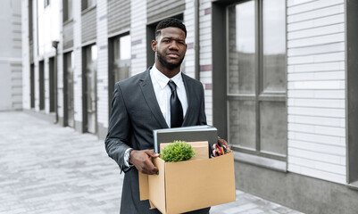 A man in a suit walks outside holding a cardboard box filled with office supplies and a small...