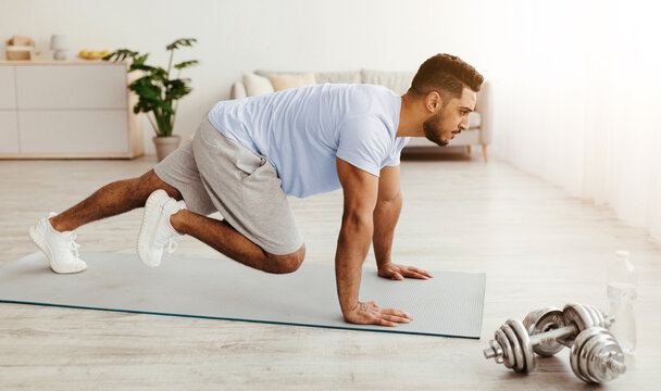 A man performs a mountain climber exercise on a yoga mat in a bright indoor space. He focuses on his fitness routine while dumbbells rest nearby.