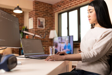 Asian software developer working remotely from home, programming across device screens. Female IT specialist seated at desk, reviewing coding scripts and maintaining online security.